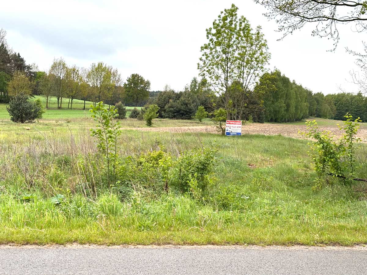 587945473_2764824190530517_4294217633839334399_n Widok na zieloną działkę z tablicą ogłoszeniową "Na sprzedaż" umieszczoną wśród drzew i łąk.
