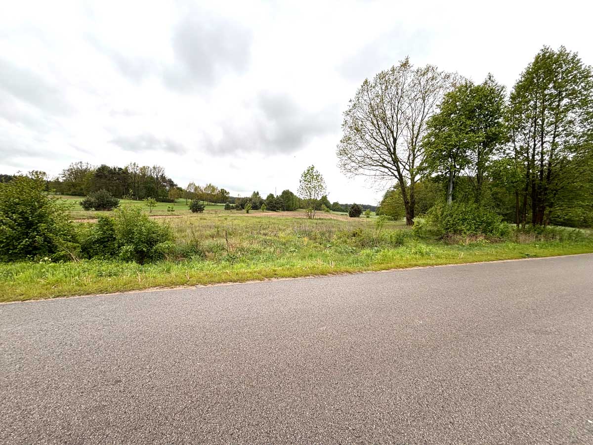 587959700_2764824117197191_8703529935299917411_n Widok na wiejską drogę otoczoną zielonymi łąkami i drzewami w pochmurny dzień.