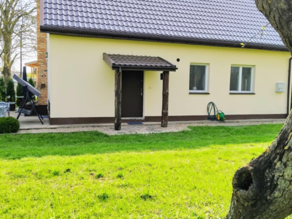 Dom jednorodzinny z jasną elewacją i ciemnym dachem, otoczony zielonym trawnikiem i drzewami. W tle widoczny taras oraz sprzęt ogrodowy.