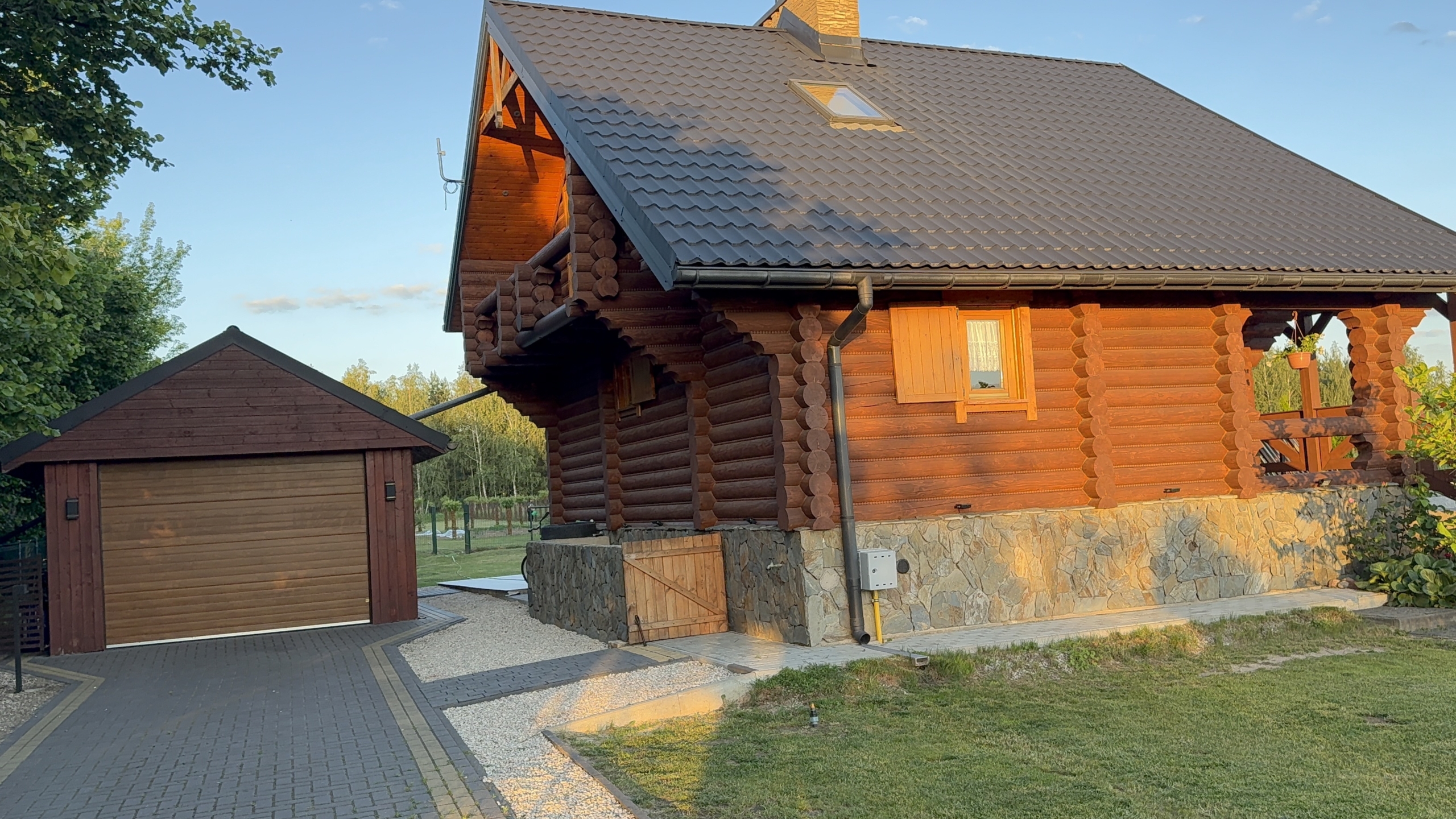 Drewniany dom z tarasem i garażem, otoczony zielenią, z płaskim dachem pokrytym dachówką.