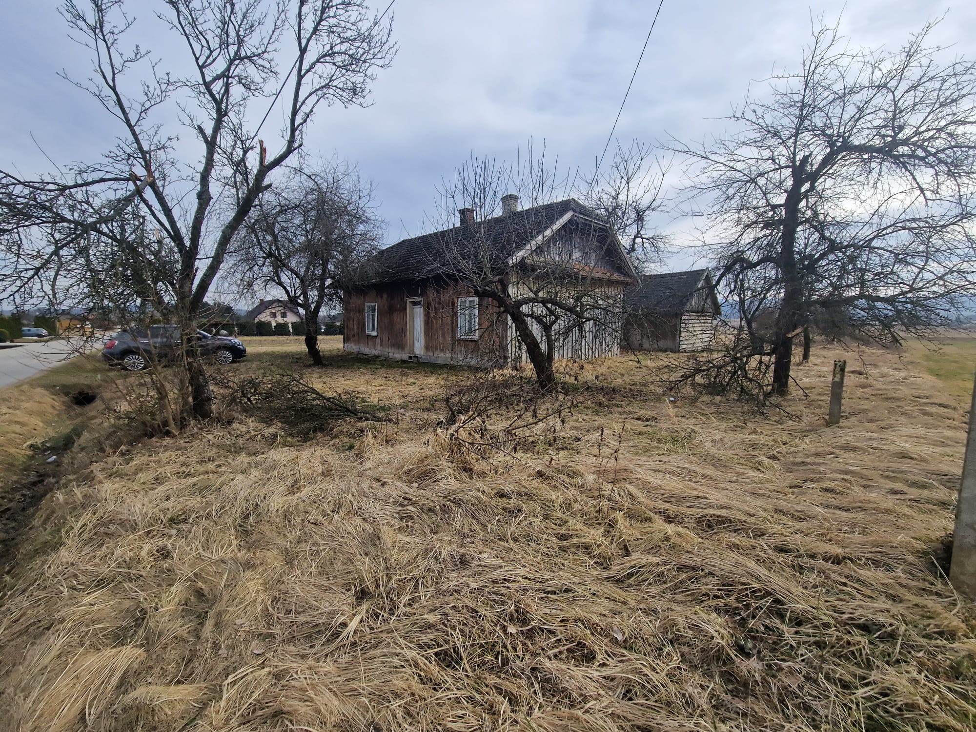 Stary dom na wsi otoczony nagimi drzewami i trawą, z widocznym samochodem zaparkowanym przy drodze.